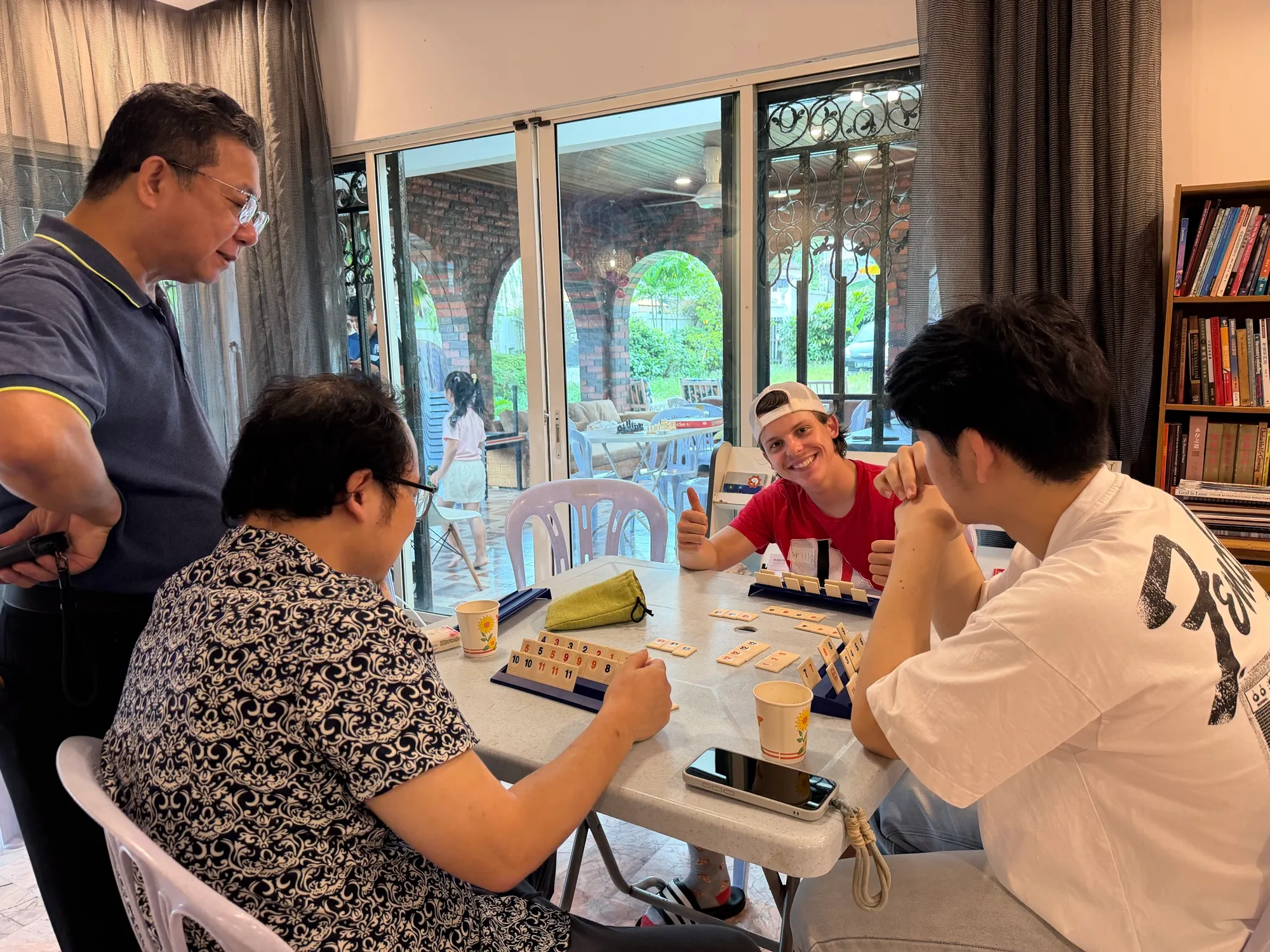 Students and volunteers playing a tile game around a table, one of them giving a thumbs up