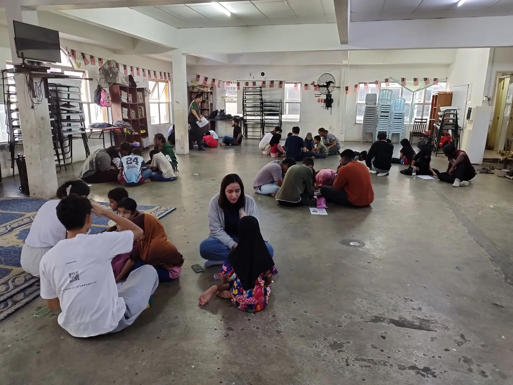 University student volunteers sitting on the floor in small groups with children at a refugee learning centre