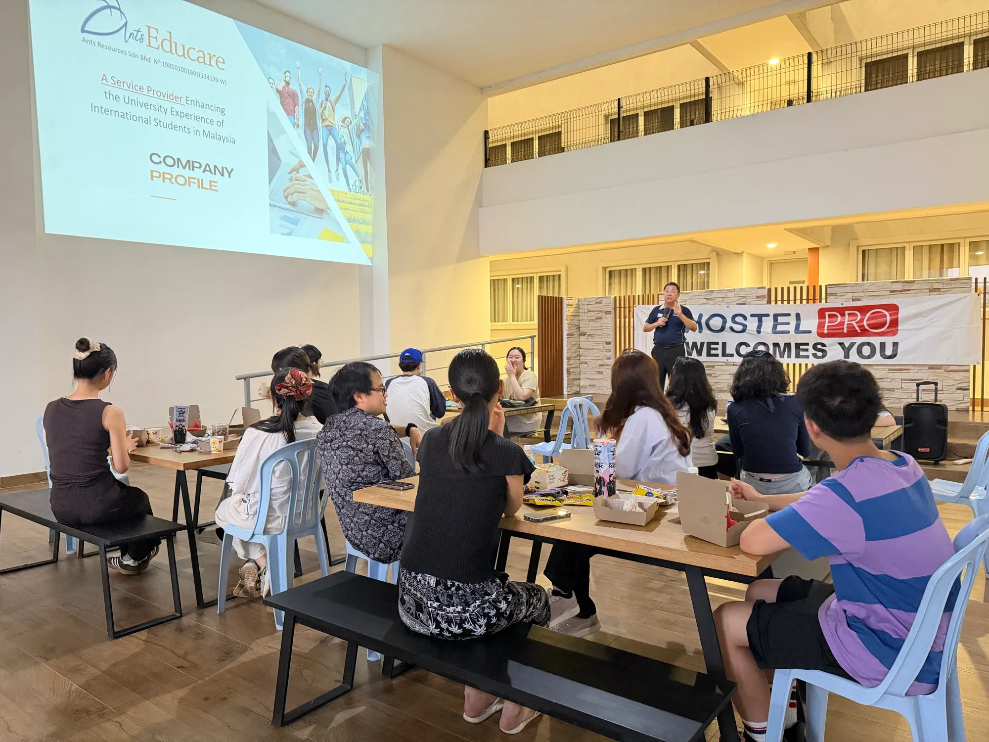 International students at a welcome dinner hosted with HostelPro, listening to a speaker in front of a HostelPro banner